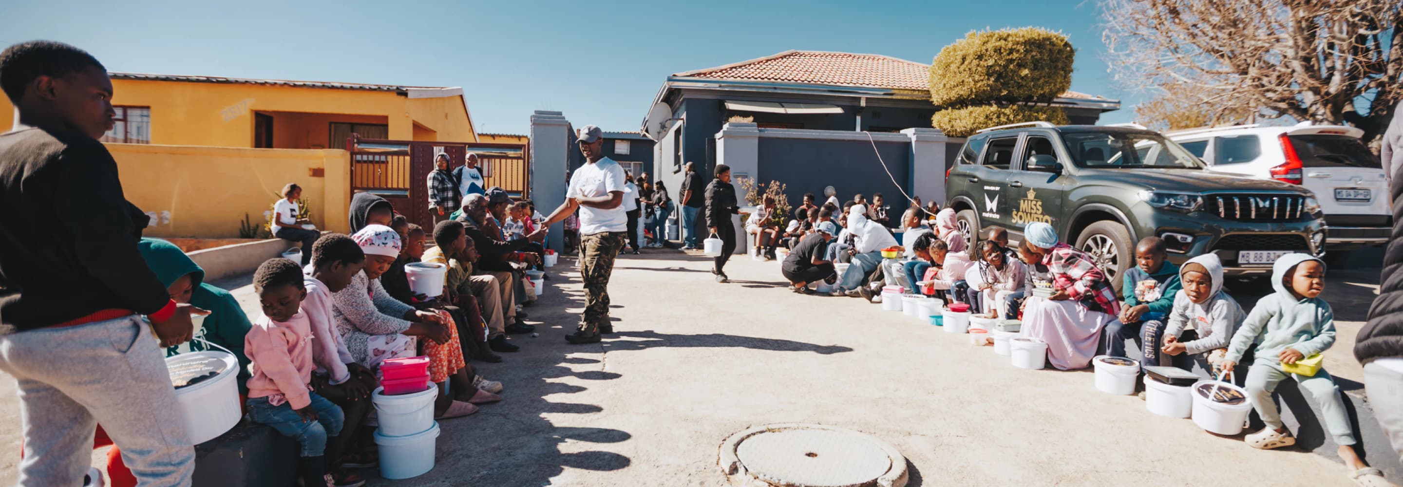 Community members seated outdoors beside a Mahindra Scorpio-N during Mandela Day distribution