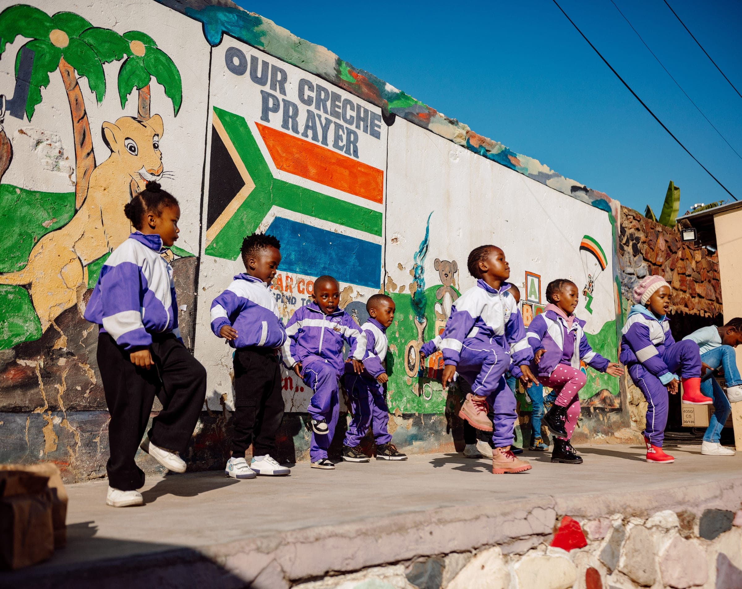 Children in purple tracksuits dancing in front of the Ba-Tliseng mural wall